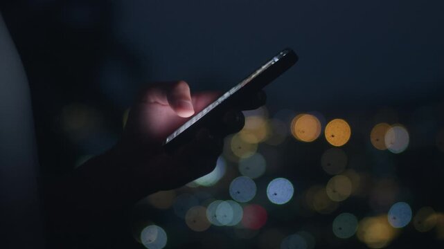 Smartphone in hand is used at night while a male hand scrolls social media against blurred city lights. Smartphone in hand reflects mobile addiction urban solitude and nonstop digital communication.