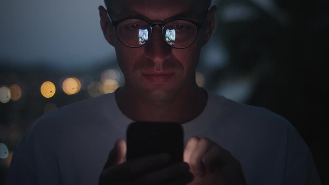 Close up of man scrolling social media on smartphone at night with glowing screen in glasses. Concept of smartphone dependence digital lifestyle modern communication and nonstop online presence.