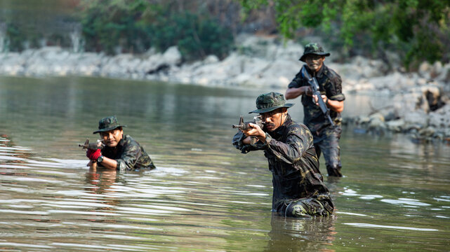 Camouflaged Soldiers Conducting Tactical Operations Near Murky River Shoreline, Wearing Dark Camouflage Clothing and Protective Eyewear, Engaging in Intense Combat Training.