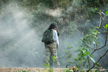 Naklejka premium Woman in Camouflage Fatigues Conducting Forest Terrain Inspection with Backpack and Blurred Observer in Dense, Misty Green Woods, Assessing Landscape Details.