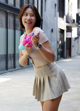 A happy asian woman stands in an urban alleyway holding a colorful water gun and laughing during a playful moment