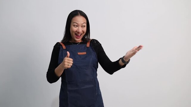 A cheerful Asian female barista with blue apron presenting aside enthusiastically with a big smile while making thumbs up
