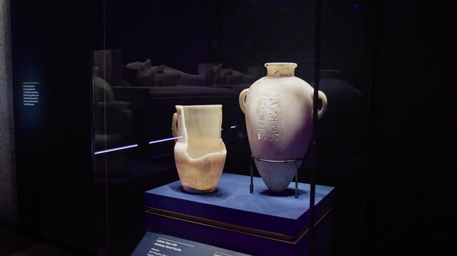 Ancient Egyptian alabaster vessels, including a canopic jar, displayed in a dimly lit museum case with inscriptions and preservation mounts.