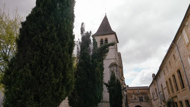 A stone church spire rises above tall cypress trees in the medieval village of Monpazier, Dordogne. Slow motion traveling movement along buildings.