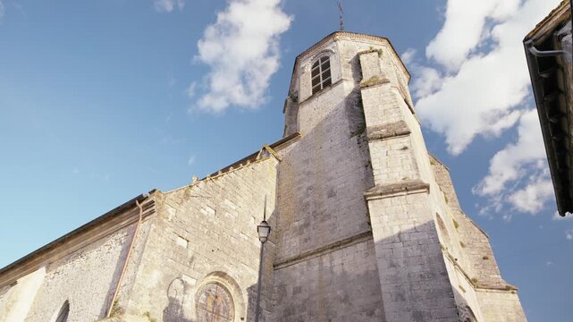 Low-angle view of the bell tower and stone structure of the church in the village of Issigeac. Historic monument under a blue sky. Circular slow motion tracking shot.