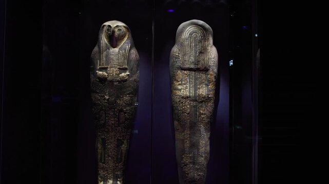Front and rear views of an ancient Egyptian sarcophagus with falcon mask, richly decorated with hieroglyphs, displayed in dark museum curation lighting.