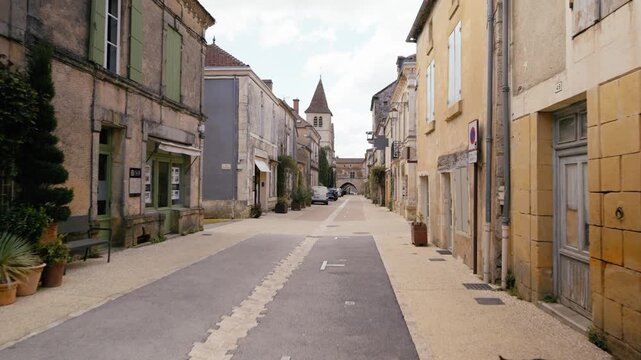 Walking through a narrow paved street in Monpazier, Dordogne, featuring traditional stone architecture and a church tower. Slow motion tracking shot.