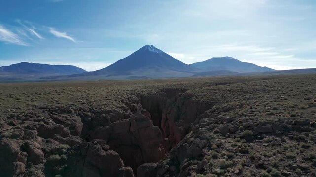 Backward drone flight over a dramatic rocky ravine revealing a vast arid altiplano plateau with Licancabur snow capped volcano dominating the horizon. San Pedro de Atacama, Chile