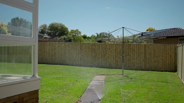 Static shot of a suburban residential backyard with a sprinkler actively watering
a green lawn on a bright sunny day. A classic Hill Hoist rotary clothesline stands
in the yard.