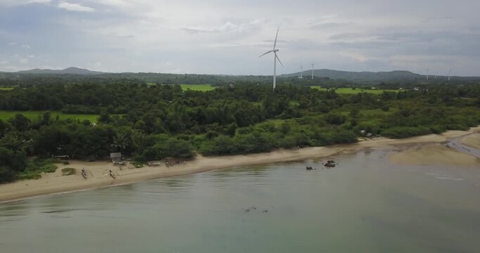 Aerial drone shot over calm sea moving slowly toward wind turbine on tropical coastline with green landscape in Guimaras Philippines