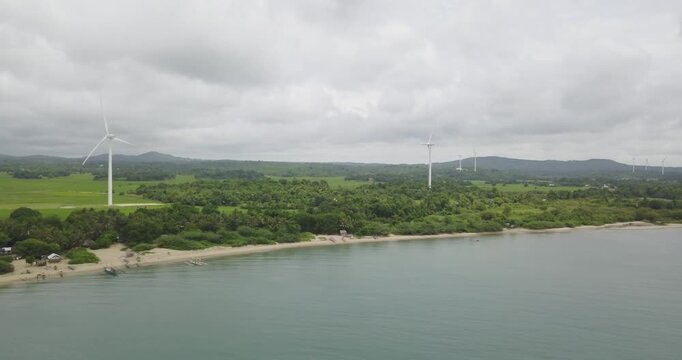 Aerial shot moving forward from calm ocean toward tropical coastline with multiple wind turbines generating renewable energy across lush rural landscape in Guimaras Philippines
