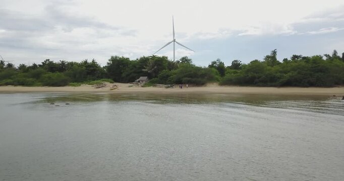 Low altitude aerial shot moving forward from calm water toward tropical beach with centered wind turbine generating renewable energy in lush coastal landscape in Guimaras Philippines
