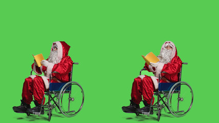 Naklejka premium Santa in wheelchair reads book, enjoying lecture novel story over greenscreen full body. Young man dressed as saint nick with disability, reading literature or fiction for knowledge.