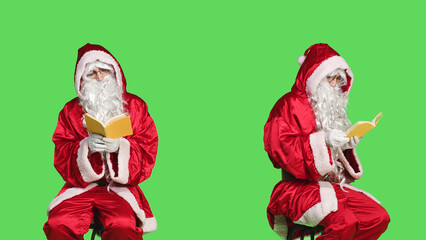 Naklejka premium Santa reads literature sitting on chair, man wearing festive red costume while he is reading poetry book over greenscreen backdrop. Father christmas enjoying fairytale story, winter holiday.