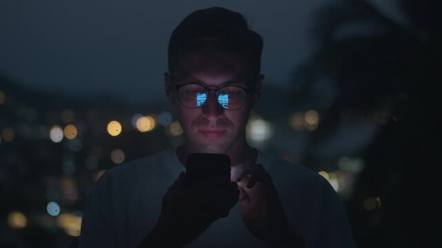 Man checking mobile phone at night scrolling social media with screen reflection in glasses. Close up portrait shows mobile phone habit digital lifestyle technology use and online communication.
