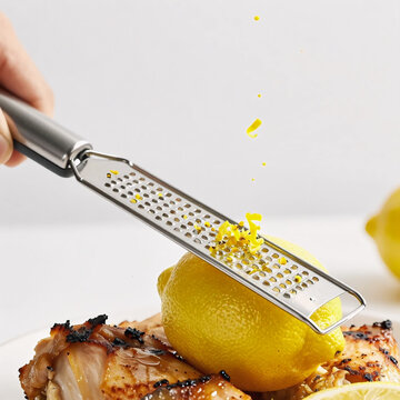 practical kitchen microplane tool grating yellow lemon peel over cooked roasted chicken,
handheld zester zesting a halved lemon onto a plate with grilled chicken and lemon slices.