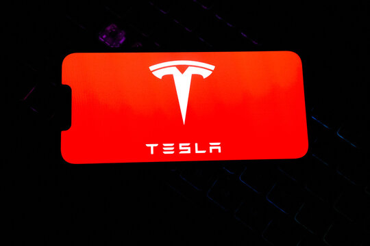 White Tesla Logo and Brand Name Displayed on a Bright Red Smartphone Screen Over a Dark Backlit Mechanical Gaming Keyboard Background - Concept for Electric Vehicles, Tech Innovation, and Clean Energy
