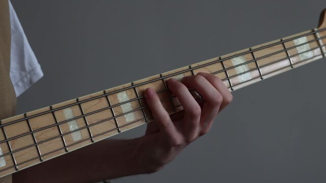 Close up of a person is playing notes on the fretboard of a bass guitar, the background is a neutral grey colour and you can see the individual notes being played