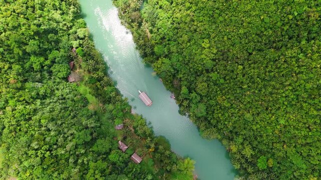Aerial top-down view of a cruise boat navigating the turquoise Loboc River in Bohol. Tropical landscape with lush green jungle palm trees surrounding the water. Exotic travel destination