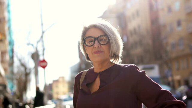 Elegant mature woman with stylish grey hair and glasses standing confidently on a sunny city street, looking at the camera with a slight smile and her arms crossed on a beautiful day