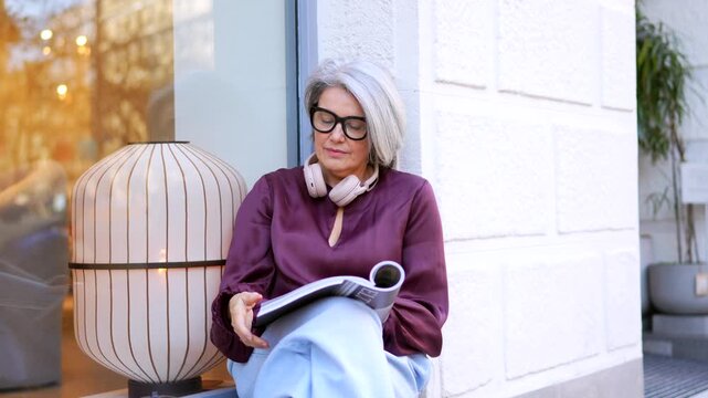 Stylish senior woman with grey hair and glasses sitting on a windowsill in the city, relaxing while reading a magazine and enjoying a moment of leisure with a coffee cup nearby