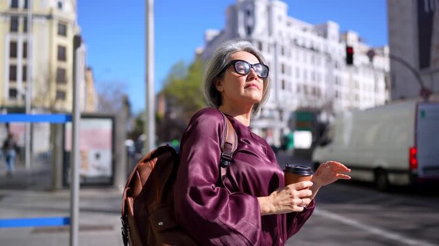 Elegant senior woman with grey hair and sunglasses waiting at a bus stop, then raising her hand to hail a taxi cab on a sunny city street while holding a cup of coffee and a backpack