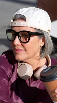 Stylish mature woman with gray hair wearing a backward baseball cap and eyeglasses, smiling and drinking coffee from a paper cup while enjoying a sunny day outdoors in the city