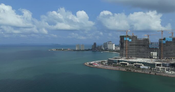 Aerial view of massive coastal skyscrapers under construction in Sihanoukville with numerous cranes and a distant peninsula city skyline in the background.