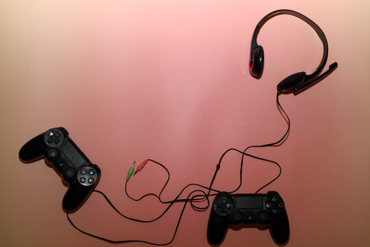 Lviv, Ukraine - March 19, 2026: Two black gaming controllers and a wired headset with a microphone lie randomly on a soft pink background.
