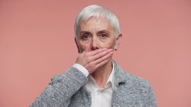 Senior woman covering mouth in silence gesture on pink