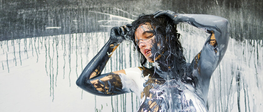 Sexy nude woman covered in abstract black, white, and gold paint, standing against a dripping, textured backdrop, embodying a fusion of art and human form