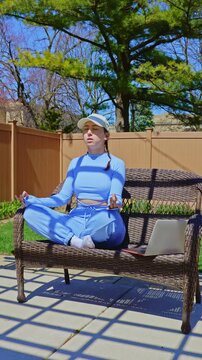A woman sits cross-legged on a garden bench while meditating. She has a laptop next to her and is in a park with green trees and a fence. It is sunny outside.