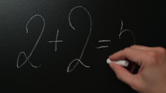 A hand shows a math equation on a chalkboard while correcting an addition mistake. The action takes place indoors, displaying problem-solving and teaching concepts