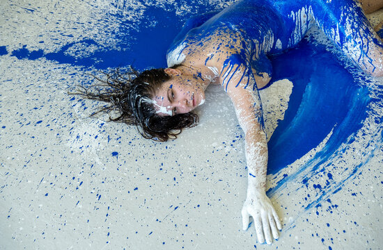 A young woman lies on the floor, her body and hair covered in bold splashes of cobalt blue and white paint. The dynamic scene merges human form with expressive color and movement