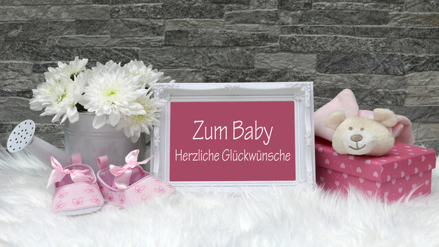 Rahmen mit der Aufschrift 'Zum Baby Herzlich Gl&uuml;ckw&uuml;nsche' auf wei&szlig;em Fell, daneben wei&szlig;e Blumen in Gie&szlig;kanne, rosa Babyschuhe und ein rosa Geschenk mit Teddyb&auml;r