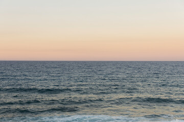 Obraz premium View of the ocean at sunset with gentle waves and a clear horizon under a pastel sky
