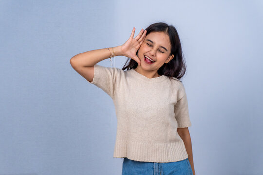 Playful Asian woman in beige shirt making a funny face and gesturing on grey background