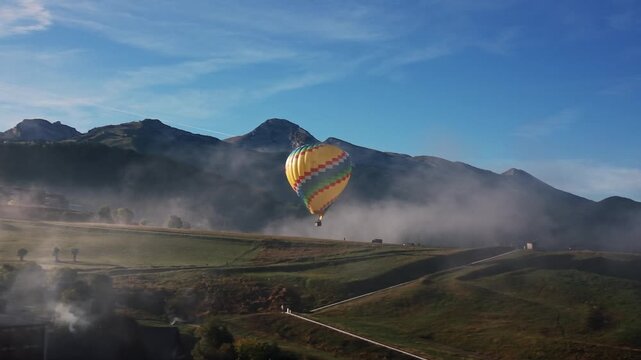 Colorful hot air balloon flying over green valley with low fog during beautiful sunrise in Auvergne-Rh&ocirc;ne-Alpes, France. Aerial drone lateral view