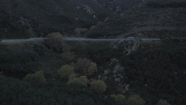 Serra da Estrela mountain road in Seia municipality Guarda district Centro region Portugal crossing rocky valley with shrub and forest biome and light vehicle traffic, aerial drone push in shot
