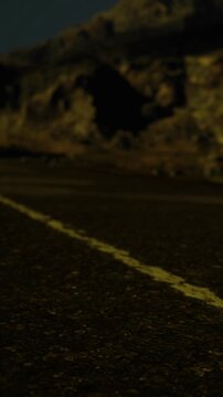 A winding road runs straight ahead under a night sky. The line on the road guides the view toward mountains in the background. The scene shows a dark landscape with a hint of light.