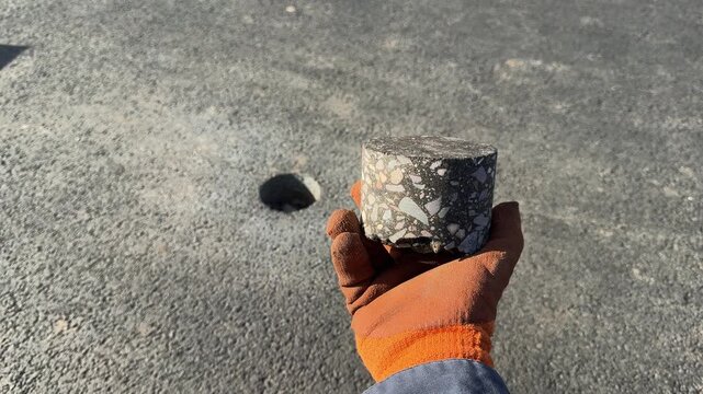Hand in protective glove holding a core sample of asphalt taken from a road surface