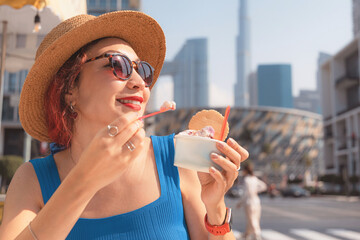 Woman smiling, experiencing a refreshing moment with gelato in front of Dubai's modern urban...