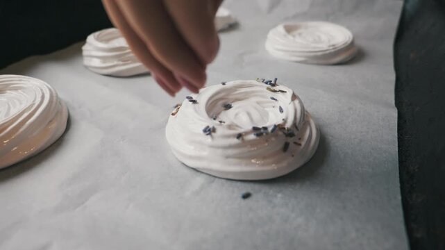 Hand sprinkles dried lavender buds over freshly piped white meringue nests on a baking sheet lined with parchment paper. Process of preparing homemade dessert treats in a kitchen setting.