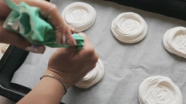 Baker hands squeeze a green pastry bag forming fluffy white meringue nests on parchment paper. The delicate dessert preparation takes place on a dark baking tray. Authentic home lifestyle.