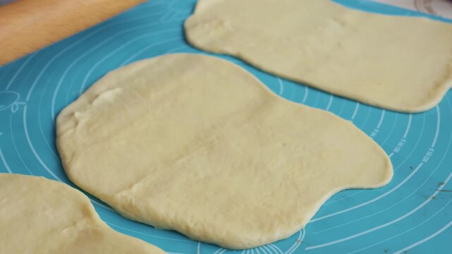 Home baker rolls out smooth yellow dough using a classic wooden rolling pin on a blue silicone mat. The hands move rhythmically to flatten the pastry for fresh muffins in a bright kitchen.