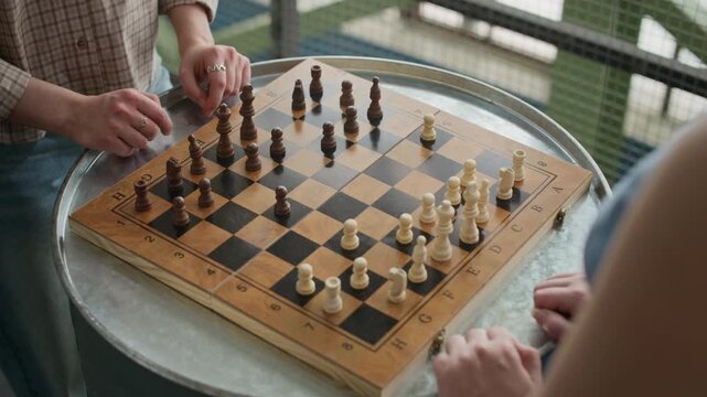 Over shoulder of two unrecognizable female players playing chess, hands moving pieces on board resting on steel keg table during focused strategic match indoors