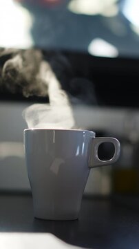 Gray ceramic mug filled with hot beverage on dark countertop, thick trail of steam rising and drifting sideways in slow motion, blurred kitchen background with bokeh light