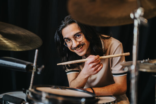A young male drummer wearing glasses smiles while holding a drumstick over his drum kit. He poses at a practice drum set in a studio setting.