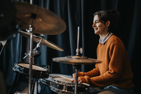 A young woman student plays a drum kit, smiling as she holds drumsticks. She rehearses on snare and cymbals in a dimly lit practice space.