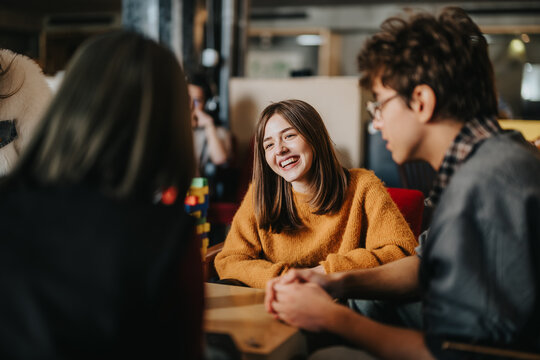 Students sit around a table smiling and talking while studying together. A casual group of young friends engages in a relaxed conversation and collaborative study session.